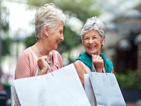 Two senior women smile at each other while carrying shopping bags